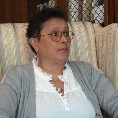 Paula Peters (Mashpee Wampanaog), wearing white blouse and grey cardigan, glasses, sits in armchair, bookcases and cabinets in background.